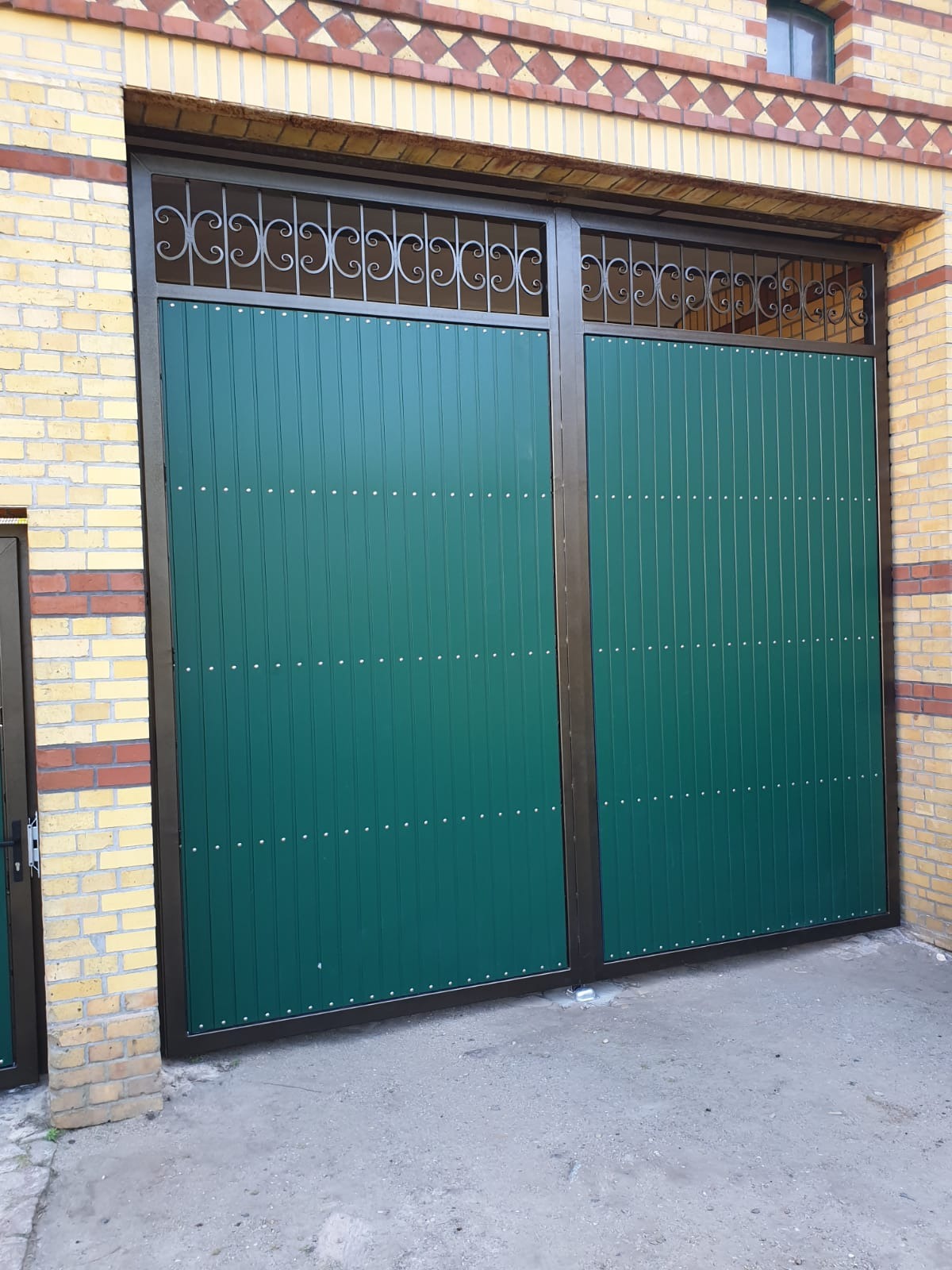 Dwuskrzydłowa, zielona brama wjazdowa z metalową, ozdobną kratą u góry, zamontowana w murze z cegły klinkierowej.