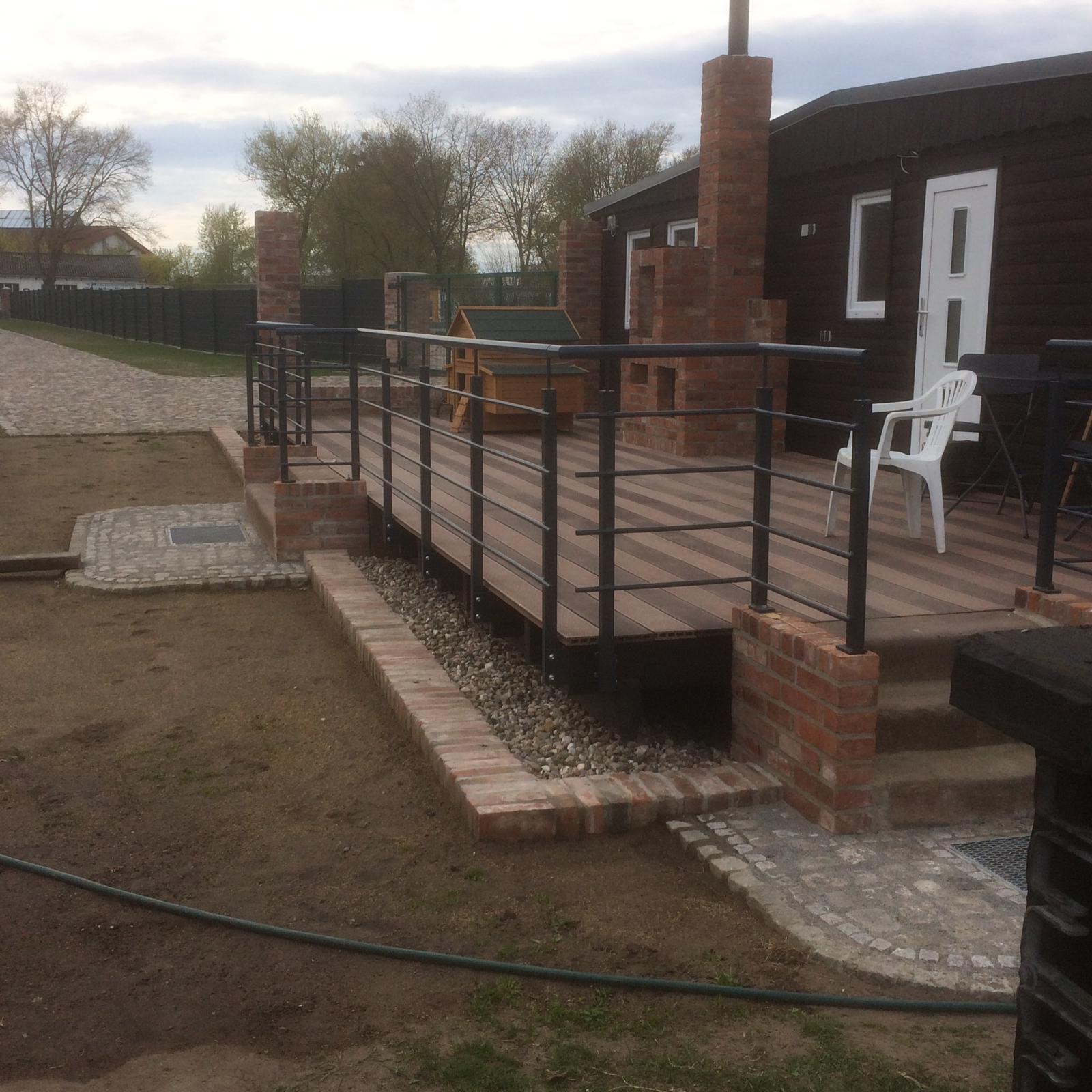 Zewnętrzny taras z drewnianą podłogą i czarną metalową balustradą, obudowany cegłą, widok na dom z ciemnego drewna i ogrodzenie w tle.