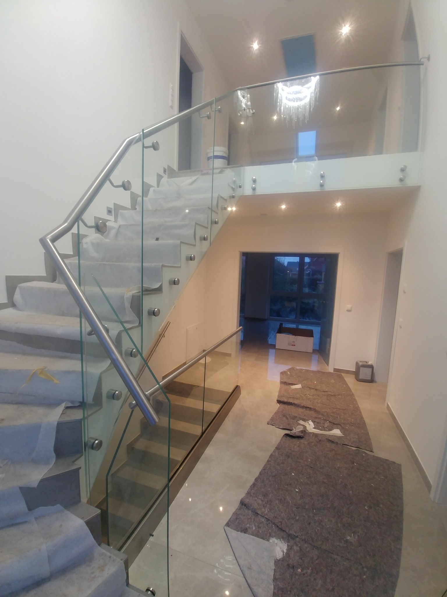 Szklana balustrada z nierdzewnymi elementami mocującymi, zabezpieczona na schodach folią ochronną podczas montażu w przestronnym holu.