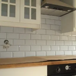 Kuchnia w trakcie remontu: białe, prostokątne płytki ułożone na ścianie, zabezpieczony blat roboczy, widoczna puszka elektryczna i fragment okapu.