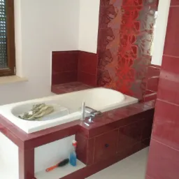 Wykończona łazienka z wanną obudowaną płytkami w odcieniach czerwieni i bieli, na półce pod wanną widoczna butelka z płynem i szczotka do czyszczenia.