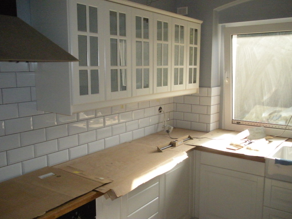 Białe, prostokątne płytki na ścianie nad blatem kuchennym, częściowo zakrytym kartonem ochronnym, pod wiszącymi szafkami z przeszklonymi frontami; widoczne narzędzia pomiarowe i gniazdko elektryczne.