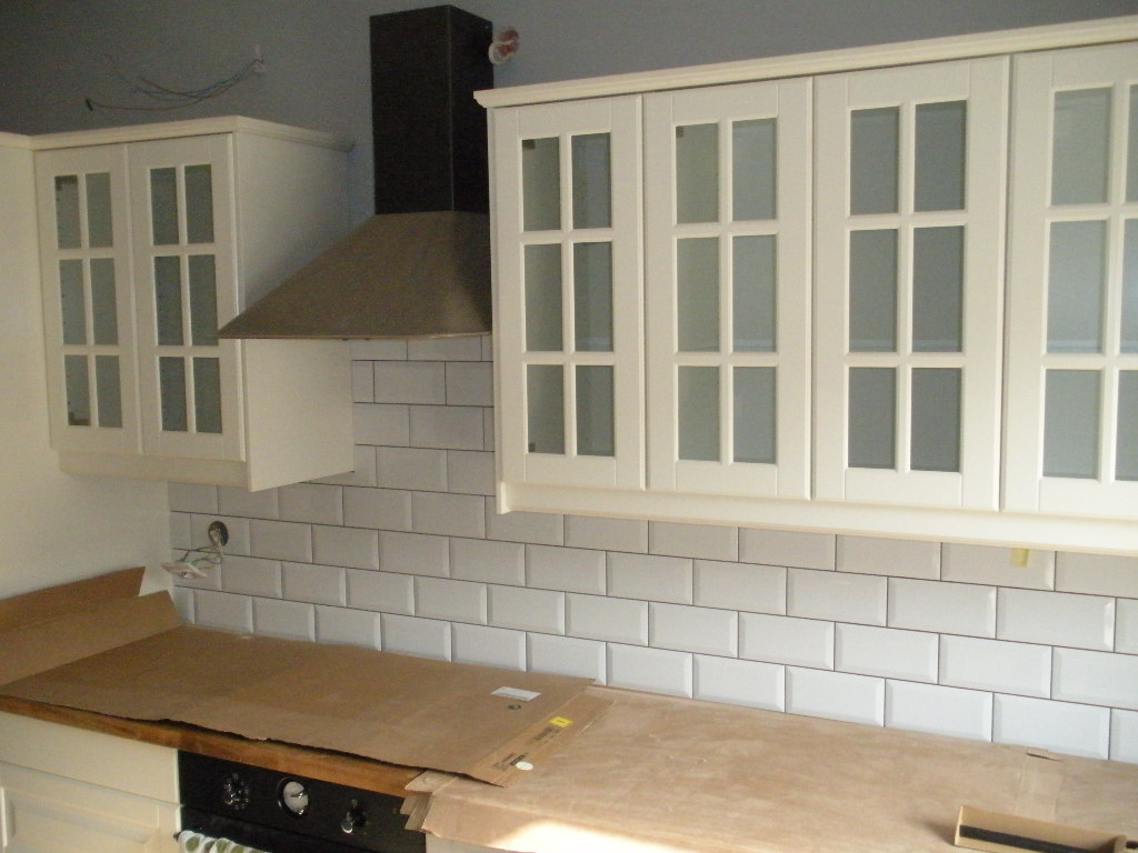 Fragment kuchni w trakcie prac wykończeniowych: białe szafki wiszące z przeszklonymi frontami, okap kuchenny, białe płytki ułożone w cegiełkę, blat roboczy zabezpieczony kartonem. Widoczne przewody...