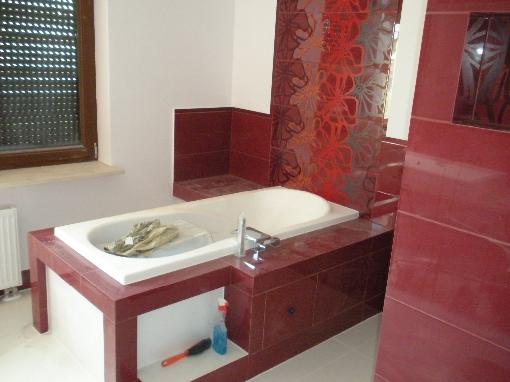 Wykończona łazienka z wanną obudowaną płytkami w odcieniach czerwieni i bieli, na półce pod wanną widoczna butelka z płynem i szczotka do czyszczenia.