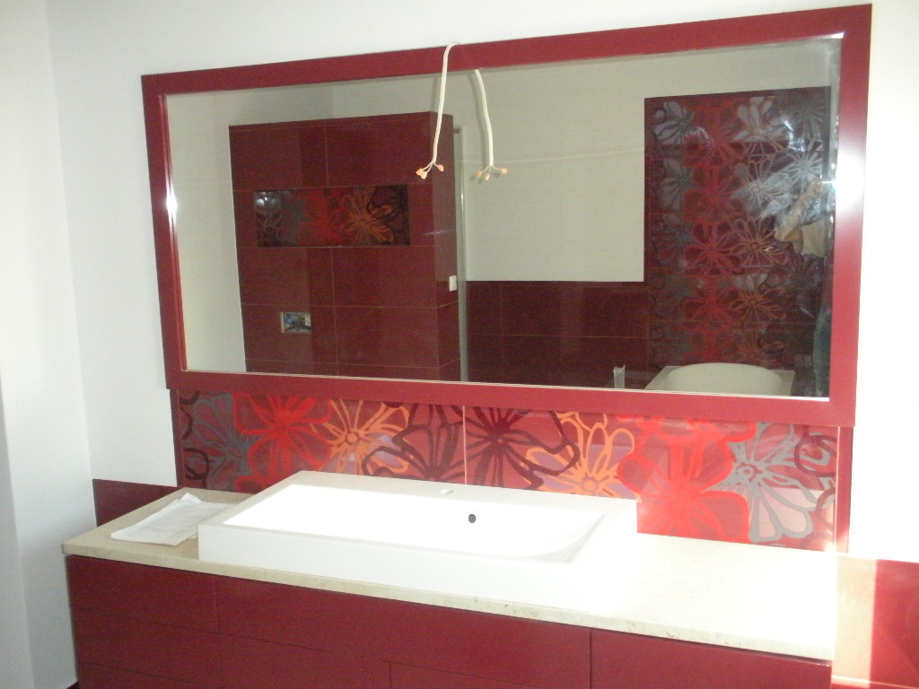 Nowoczesna łazienka w odcieniach czerwieni z umywalką wpuszczoną w blat i dużym lustrem z dekoracyjną ramą, odbijającym wykończoną ścianę z kwiatowym motywem.