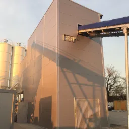 Zewnętrzny widok hali przemysłowej z płyt warstwowych, z widocznymi silosami i konstrukcją podajnika, oświetlonej popołudniowym słońcem.