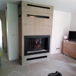 Nowoczesny kominek z obudową z betonu architektonicznego z poziomymi szczelinami wentylacyjnymi, widoczny odkurzacz warsztatowy na posadzce w trakcie prac wykończeniowych.