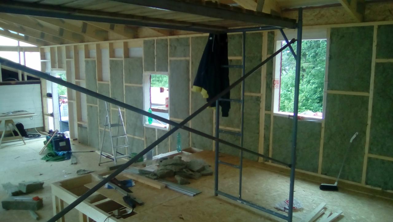 Wnętrze domu w trakcie ocieplania ścian wełną mineralną, widoczne drewniane ramy, rusztowanie, narzędzia i okno z widokiem na zieleń.