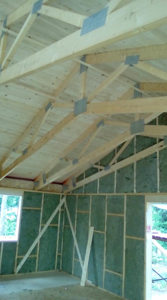 Wnętrze budynku w trakcie budowy: widoczne drewniane elementy konstrukcyjne dachu połączone metalowymi płytkami, ściany z izolacją i drewnianym szkieletem, okno z widokiem na zieleń.