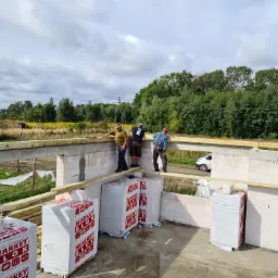 Trzech pracowników montuje drewniane belki na ścianach budynku w trakcie budowy, widoczne paczki z bloczkami komórkowymi i krajobraz za budynkiem.