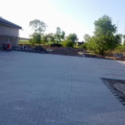 Ukończony parking z szarej kostki brukowej, widoczne wyznaczone miejsca postojowe, w tle budynek w trakcie remontu i hałda gruzu.