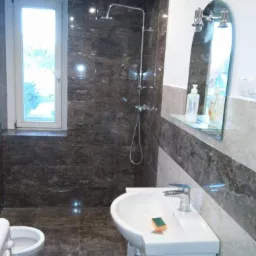 Wykończona łazienka z ciemnymi, marmurkowymi płytkami, białą umywalką z szafką i lustrem. Widoczny prysznic i okno z widokiem na zieleń.