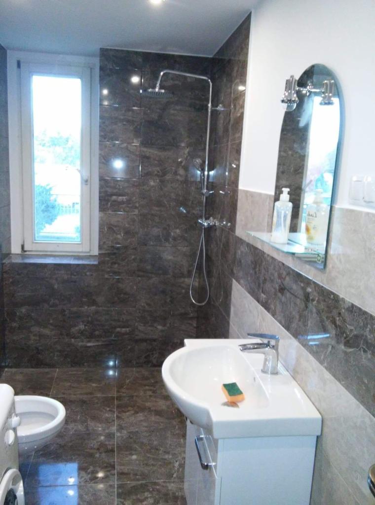 Wykończona łazienka z ciemnymi, marmurkowymi płytkami, białą umywalką z szafką i lustrem. Widoczny prysznic i okno z widokiem na zieleń.