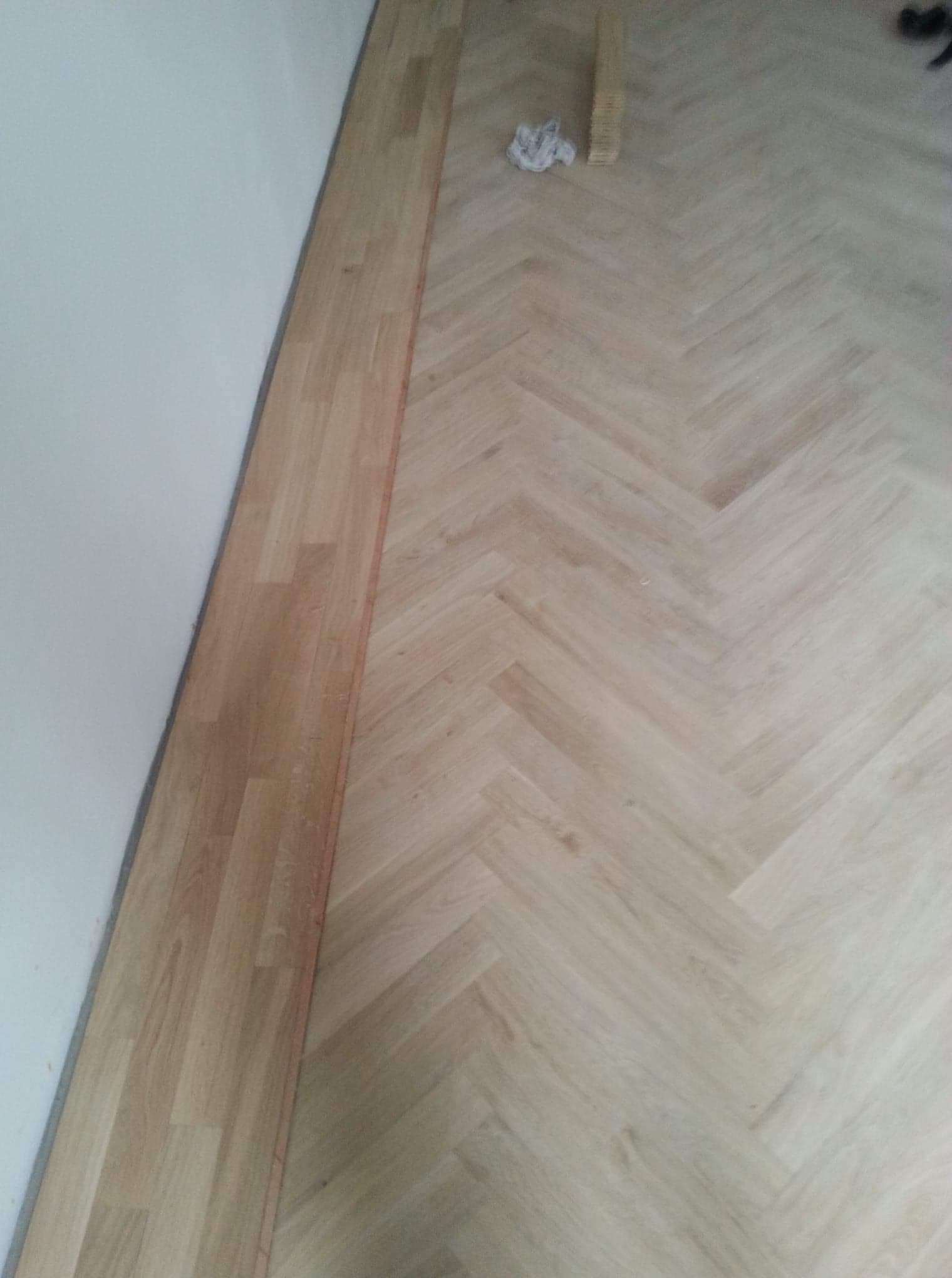 Ułożona parkietowa podłoga w jodełkę, przy ścianie listwa przypodłogowa z tego samego materiału, w rogu sterta desek i szmatka.