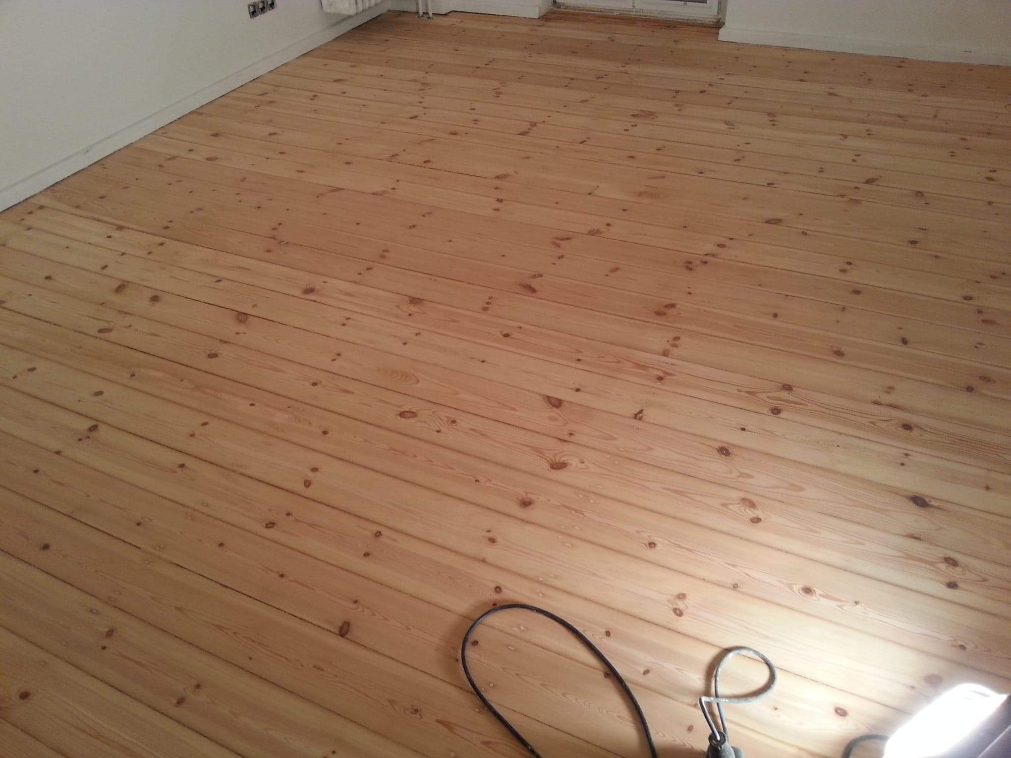 Jasna, drewniana podłoga po cyklinowaniu, widoczne sęki i naturalny rysunek drewna, w rogu kabel od szlifierki.