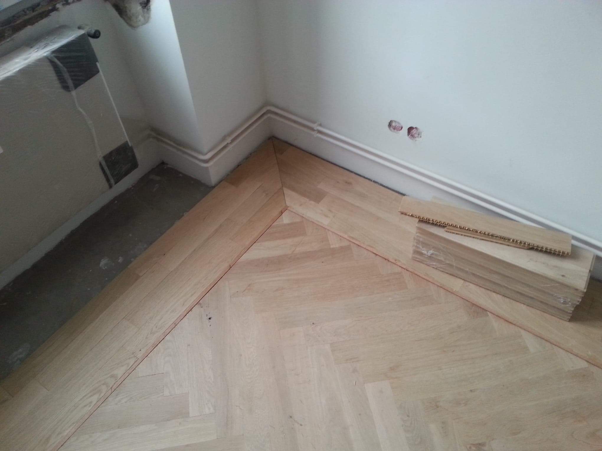 Fragment układanej podłogi z parkietu w jodełkę w rogu pomieszczenia, widoczny stos nieużytych desek, biała listwa przypodłogowa i rury grzewcze przy ścianie.
