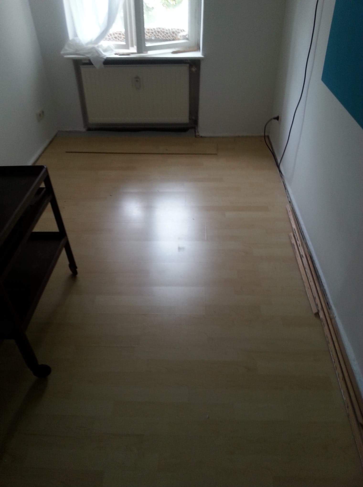 Jasna wykładzina podłogowa w pokoju z grzejnikiem pod oknem i listwami przypodłogowymi przygotowanymi do montażu przy ścianie.