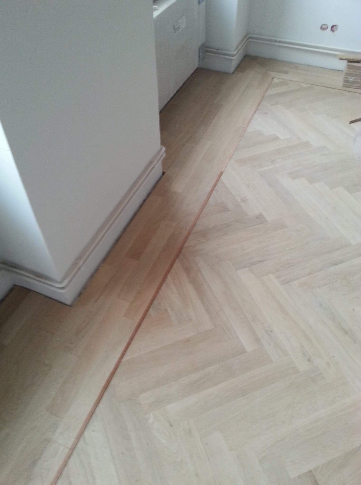 Fragment podłogi z jasnego parkietu ułożonego w jodełkę, widoczne listwy przypodłogowe przy ścianie, częściowo ułożona podłoga z widocznym fragmentem nieukończonej przestrzeni.