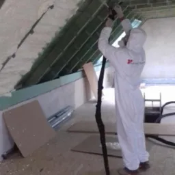 Pracownik w kombinezonie ochronnym aplikuje białą pianę izolacyjną na poddaszu, widoczne drewniane elementy konstrukcji dachu.