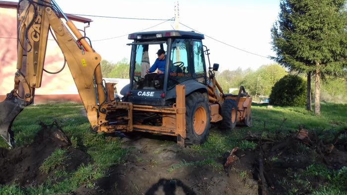 Żółto-czarna koparko-ładowarka Case w Lubieszewie podczas prac ziemnych, widok z tyłu, operator w kabinie, świeżo wykopany rów w trawie.