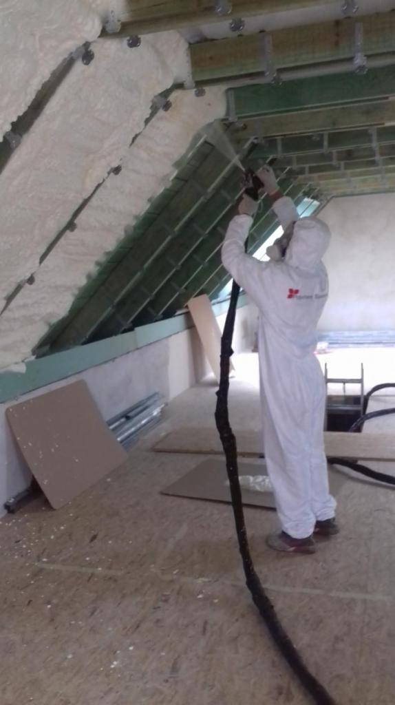 Pracownik w kombinezonie ochronnym aplikuje białą pianę izolacyjną na poddaszu, widoczne drewniane elementy konstrukcji dachu.