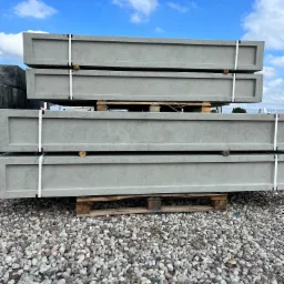 Stos czterech szarych, betonowych elementów ogrodzeniowych z wgłębieniem na górze, spiętych białymi taśmami, ułożonych na drewnianych paletach na żwirowym podłożu, z błękitnym niebem w tle.