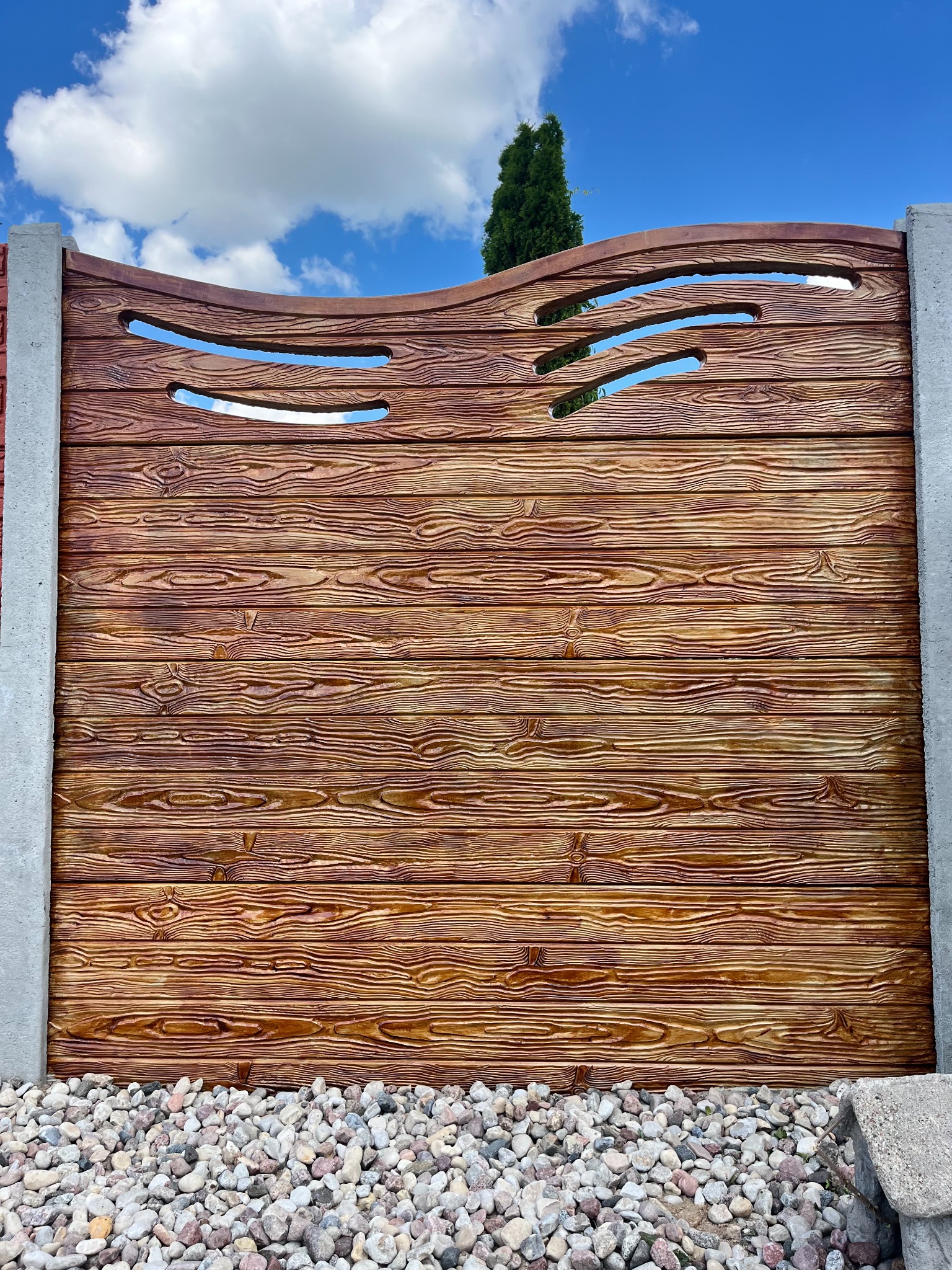 Dekoracyjny, brązowy mur z betonu imitujący drewno, z poziomymi wycięciami, osadzony na kamieniach i ograniczony szarymi słupkami, na tle błękitnego nieba z chmurami.