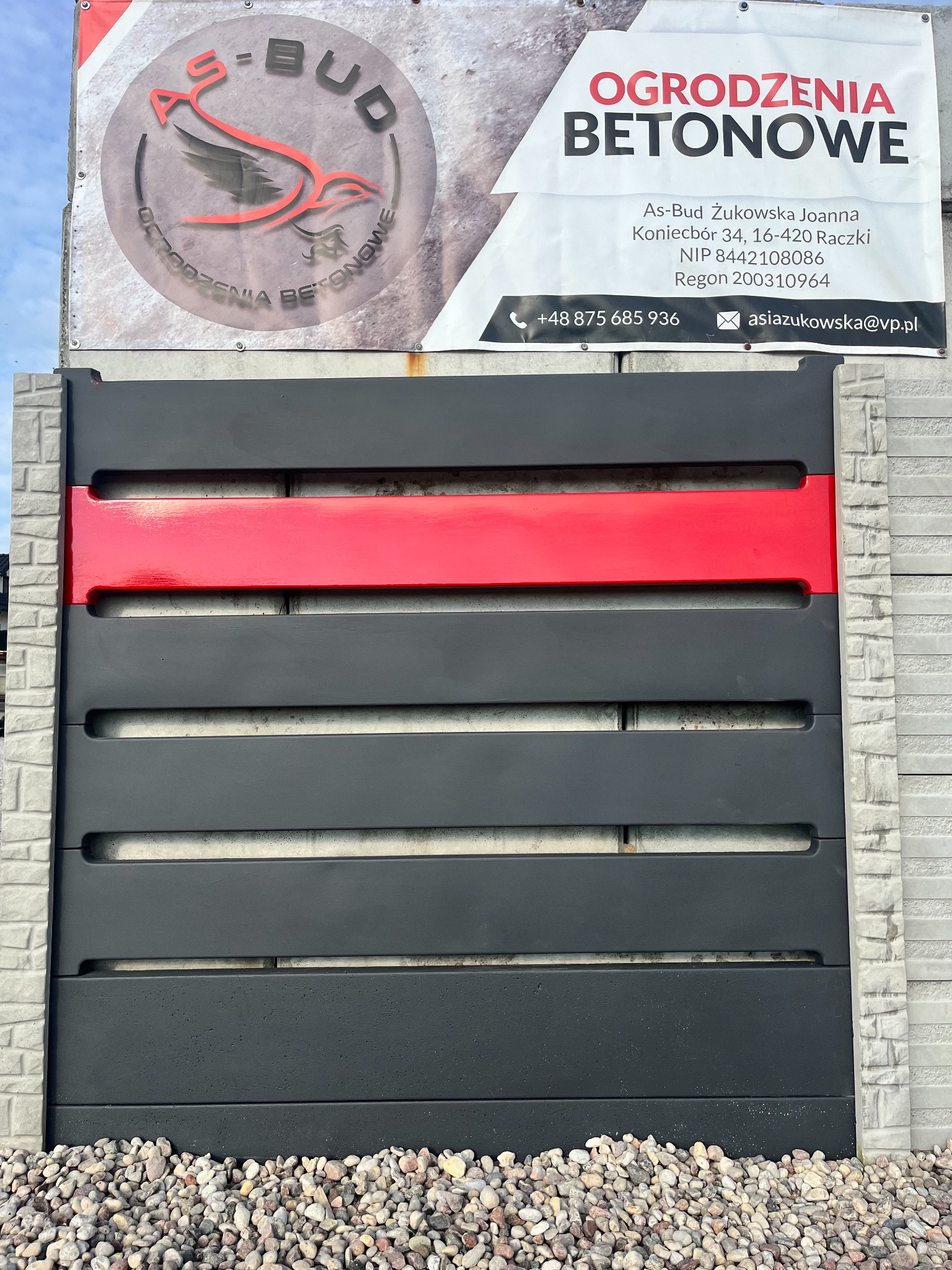 Szary panel ogrodzeniowy z poziomymi szczelinami i czerwonym pasem, na tle betonowego słupka i żwirowego podłoża, z widocznym banerem reklamowym firmy.
