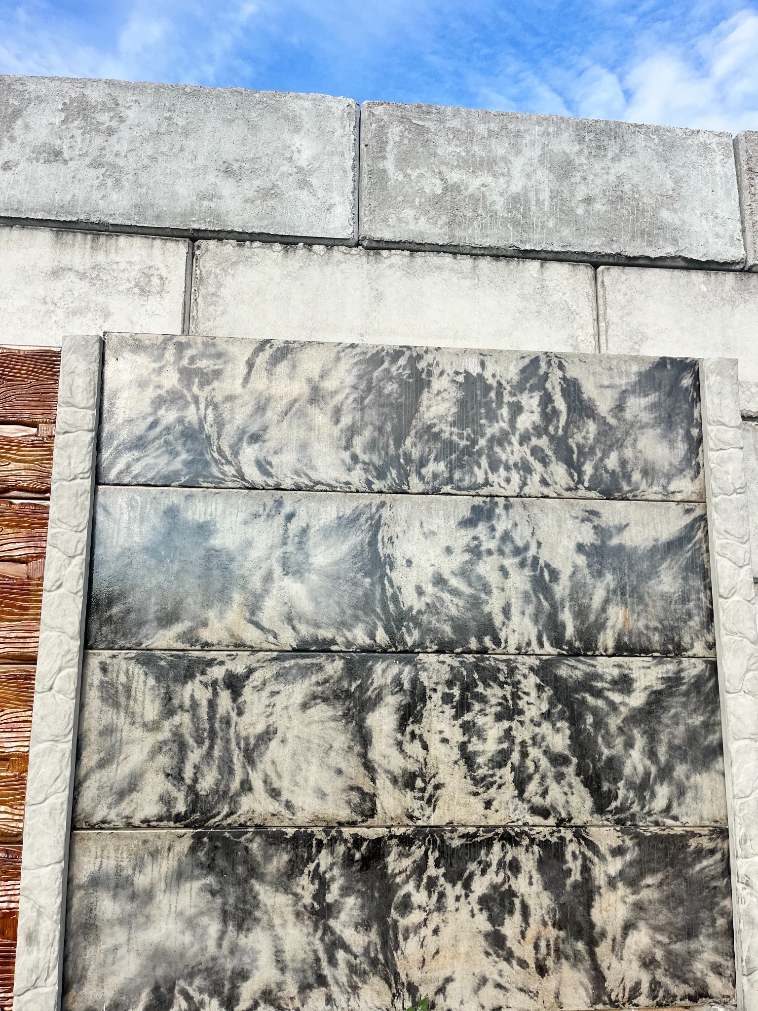 Elementy betonowe ogrodzenia: płyty z szaro-czarnym wzorem w stylu tie-dye oraz słupki z delikatnym wzorem kamienia, na tle nieba.