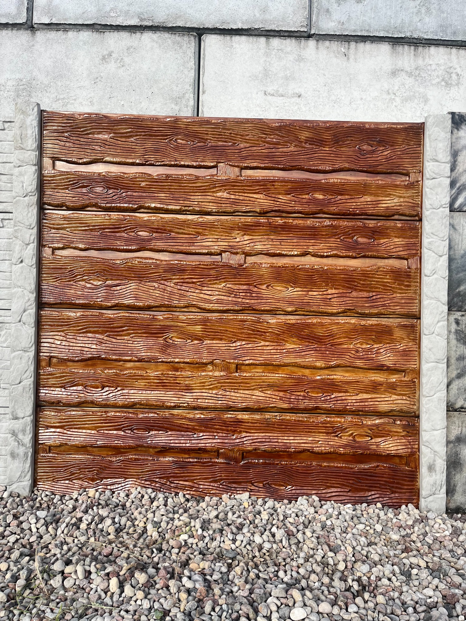 Panel ogrodzeniowy z betonu imitującego drewno, wsparty na szarych betonowych słupkach, u podstawy wysypany drobnym kamieniem.