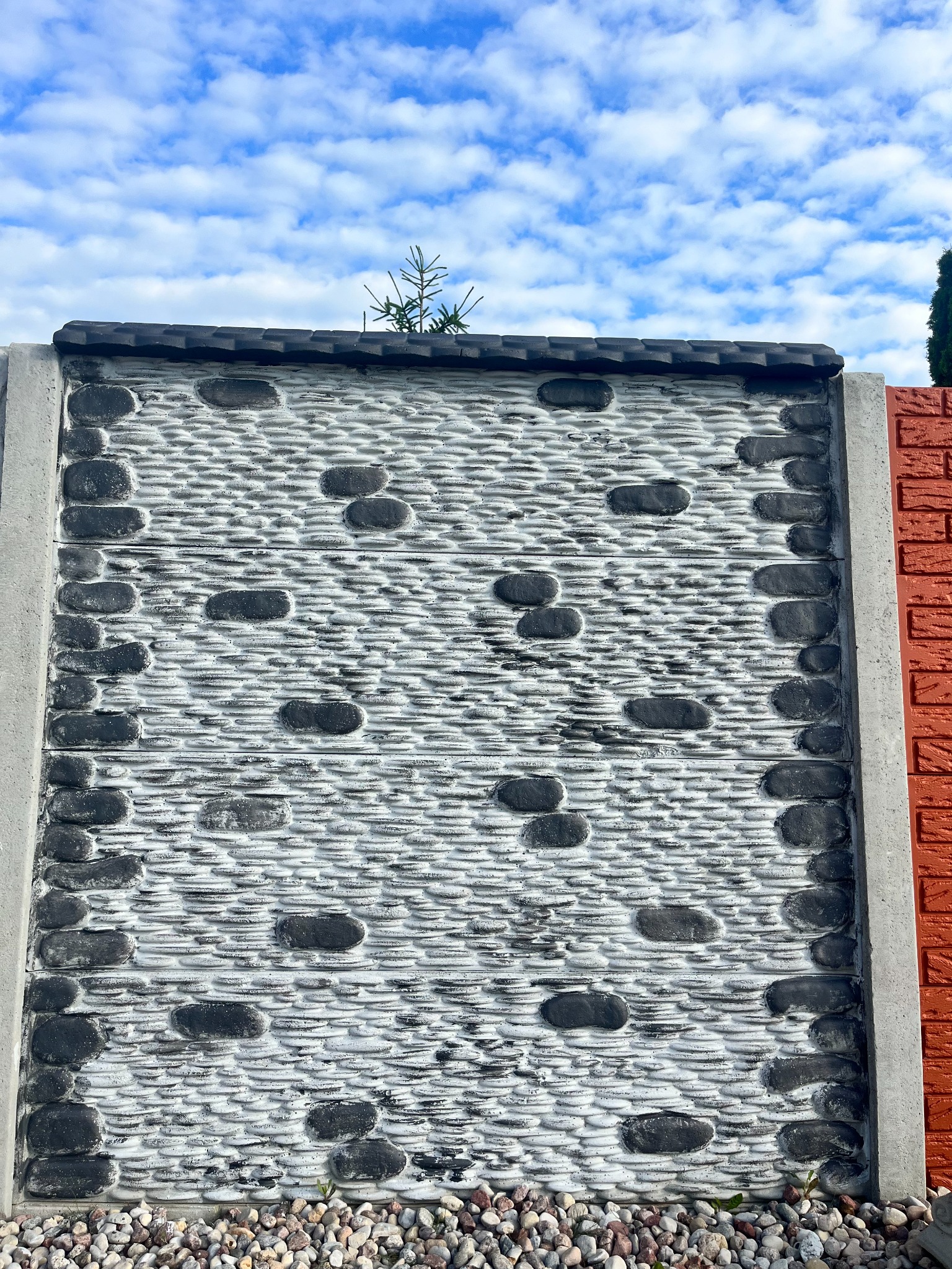 Szary prefabrykowany element ogrodzenia z fakturą imitującą kamień, obramowany betonowymi słupkami, z widocznym fragmentem muru z czerwonej cegły i niebem w tle.