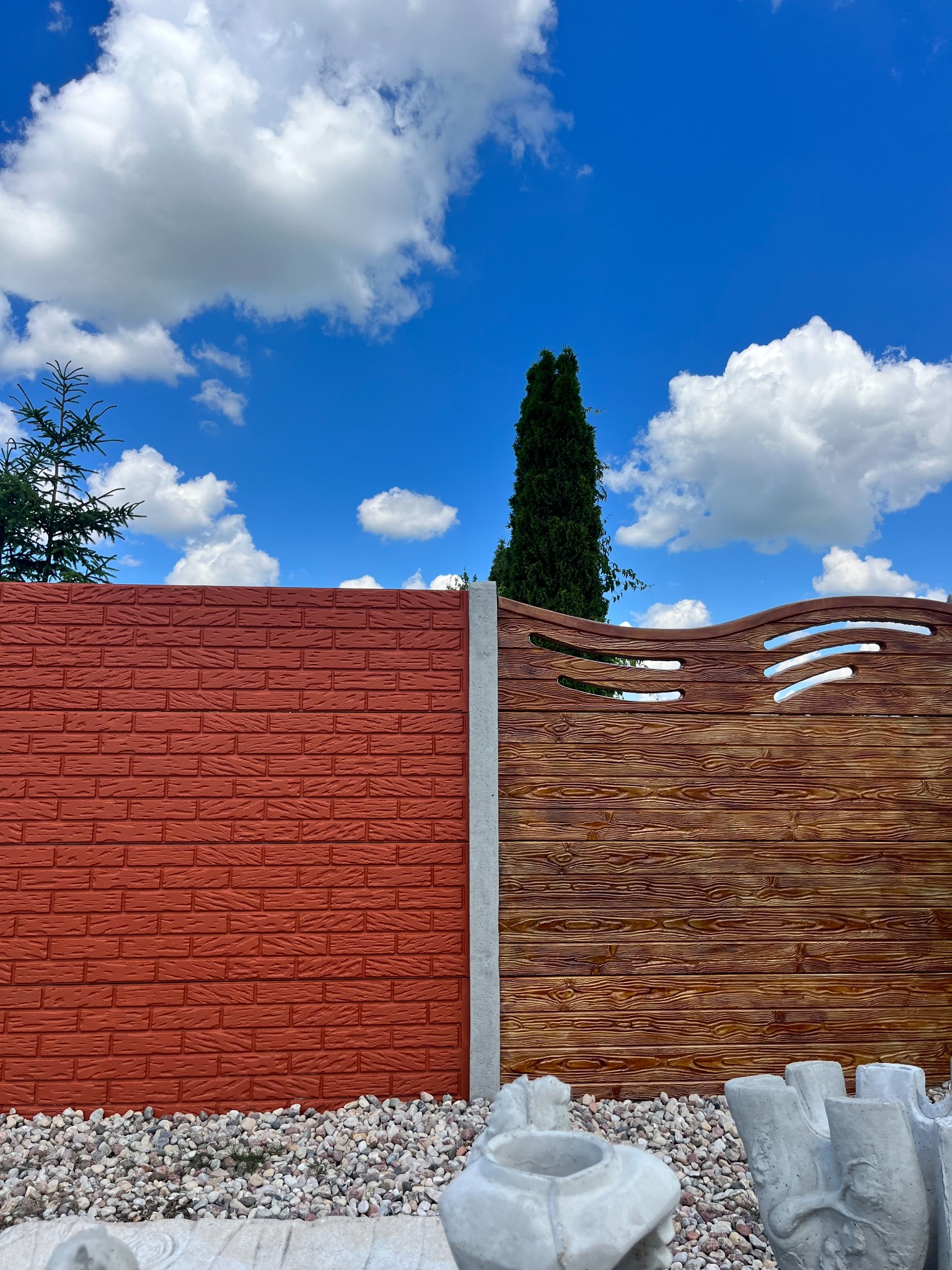 Dwukolorowe ogrodzenie z betonu imitującego cegłę i drewno, z ozdobnymi prześwitami, na tle błękitnego nieba z chmurami i drzew