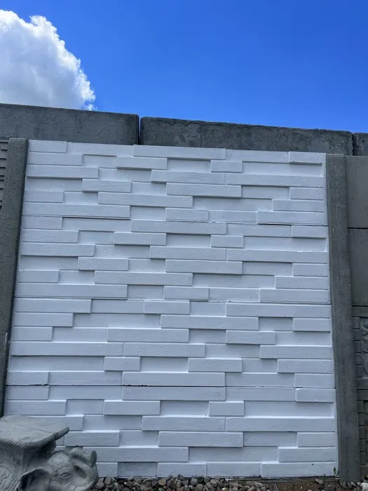 Nowoczesne ogrodzenie betonowe z białymi, poziomymi panelami o fakturze cegieł, widoczne na tle błękitnego nieba z chmurami.