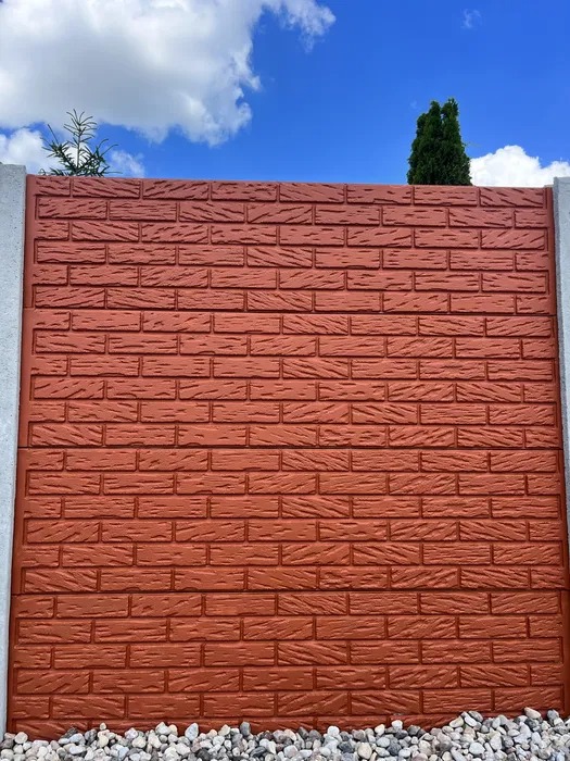 Brązowy mur imitujący cegłę z prefabrykatów betonowych, obramowany szarymi słupkami, z kamiennym podsypem i widokiem na błękitne niebo z chmurami oraz zielone drzewo w tle.