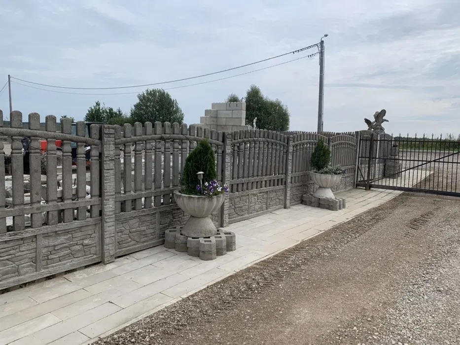 Szare ogrodzenie z prefabrykatów betonowych z dekoracyjnymi donicami i elementami imitującymi kamień, widok na posesję z żwirową drogą dojazdową.