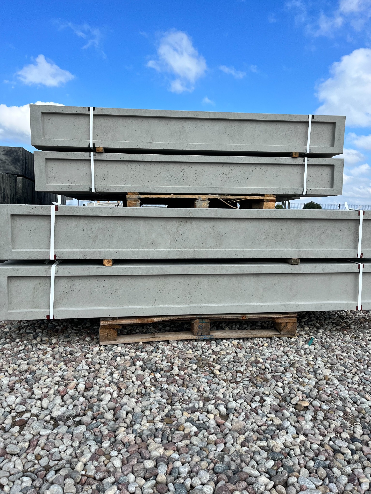 Stos czterech szarych, betonowych elementów ogrodzeniowych z wgłębieniem na górze, spiętych białymi taśmami, ułożonych na drewnianych paletach na żwirowym podłożu, z błękitnym niebem w tle.