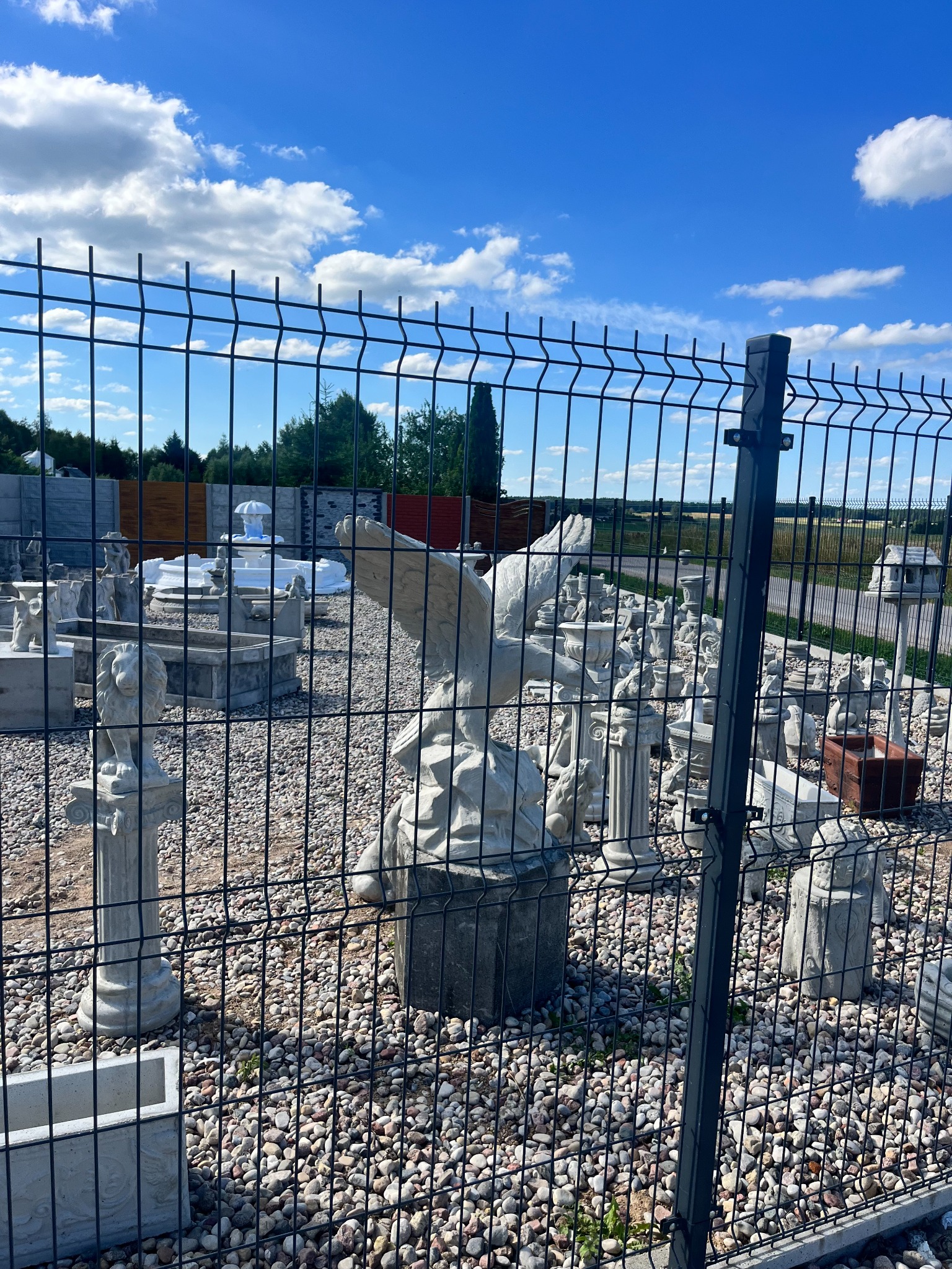 Ekspozycja różnorodnych betonowych figur ogrodowych, w tym orła, lwa i fontanny, widoczna przez metalowe panele ogrodzeniowe na żwirowym podłożu w słoneczny dzień.