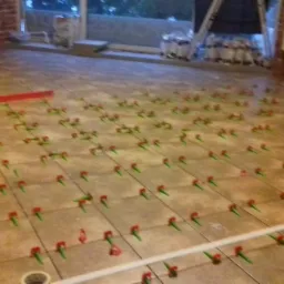 Układanie beżowych płytek podłogowych z użyciem systemu poziomowania, widoczny poziomica i worki z materiałami w tle.