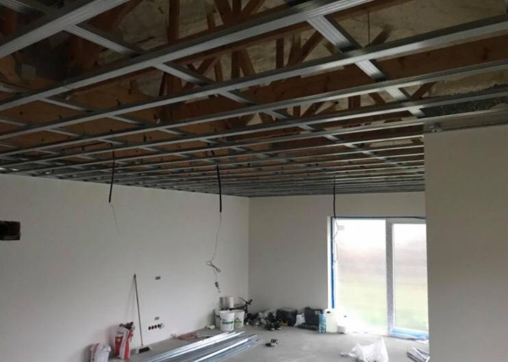 Montaż sufitu podwieszanego z widoczną konstrukcją metalową i przewodami elektrycznymi, w tle ściany przygotowane do malowania i okno balkonowe.