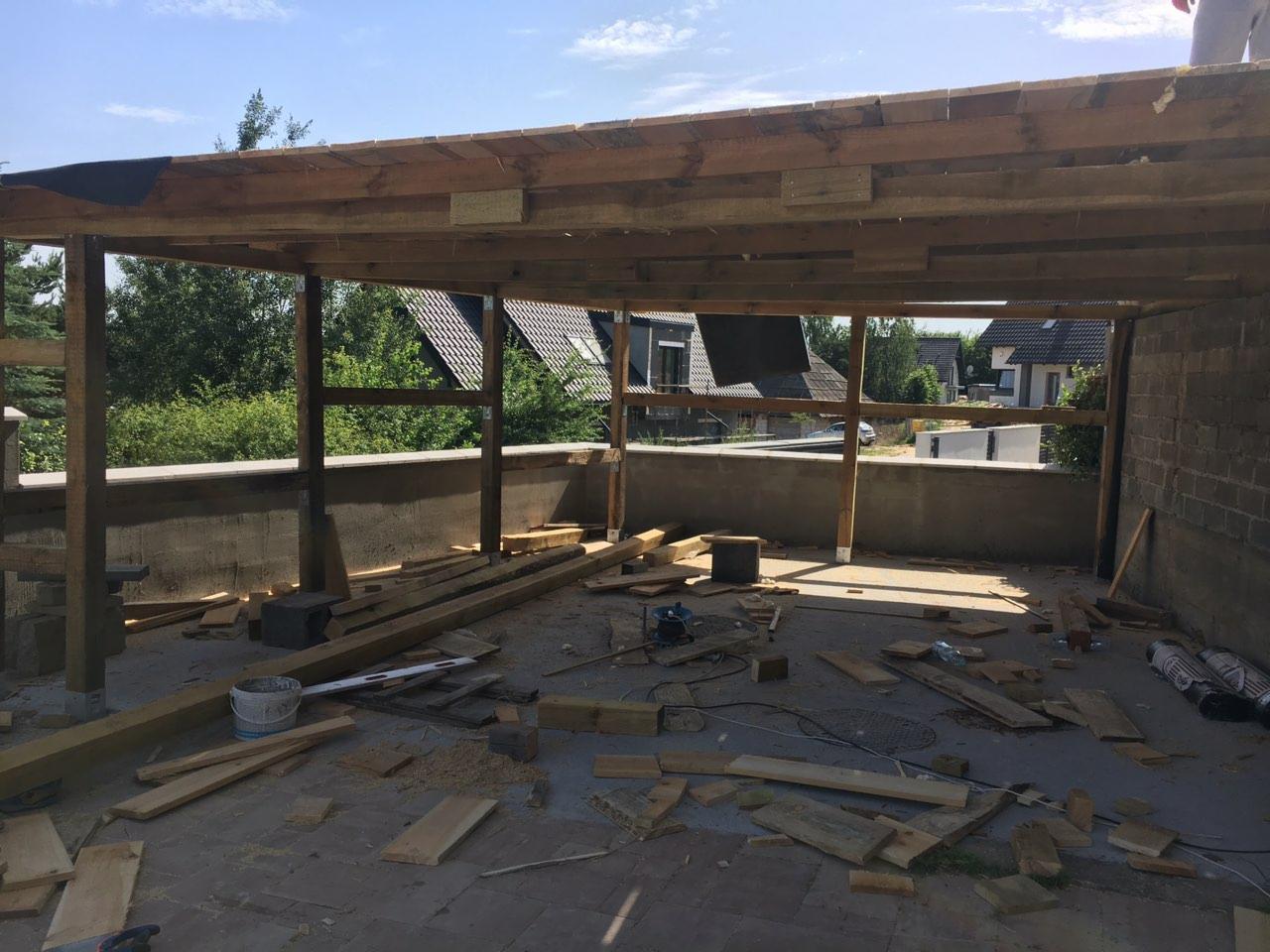 Konstrukcja drewnianego zadaszenia tarasu w trakcie budowy, widoczne deski, słupy i narzędzia na podłodze, domy w tle.