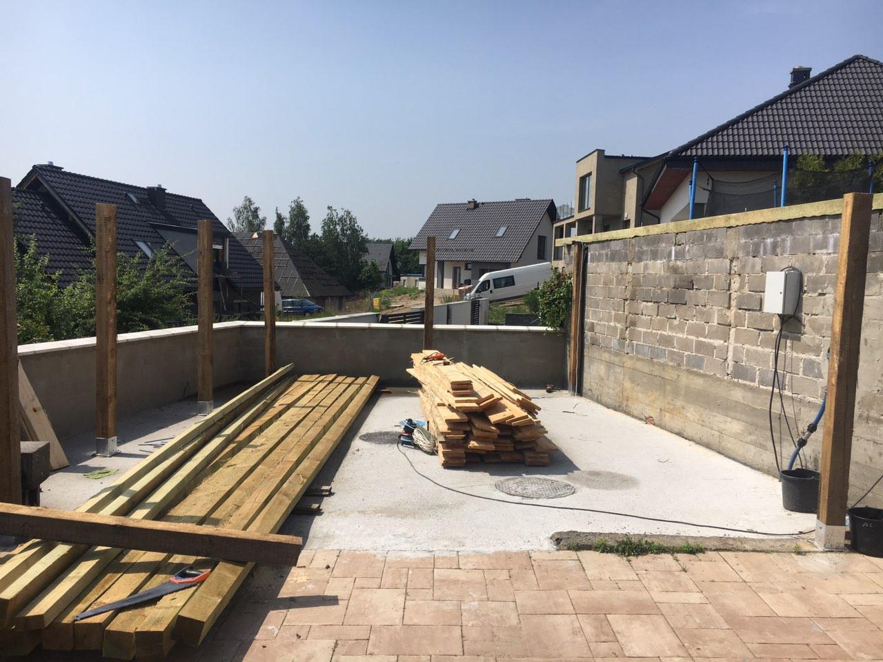 Budowa drewnianej konstrukcji na betonowym patio: ułożone deski, pionowe słupy, murowana ściana w tle, narzędzia pracy i widoczne domy w oddali.