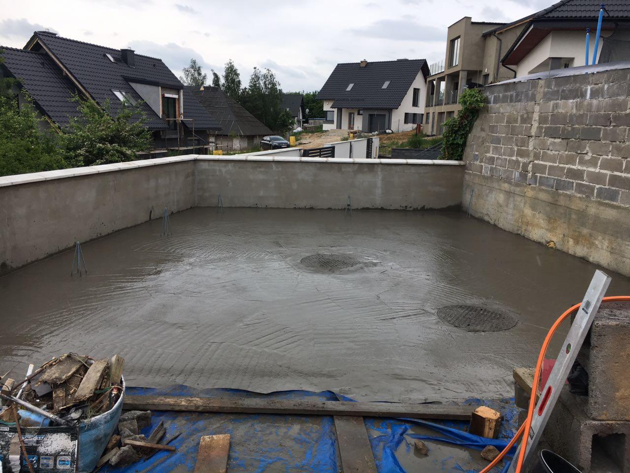 Wylewka betonowa na tarasie w trakcie realizacji, widoczne metalowe pręty, poziomica oparta o bloczki betonowe, wiadro z narzędziami i resztkami materiałów budowlanych w rogu, w tle zabudowa...