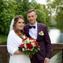 Para młoda pozuje na drewnianym moście z widokiem na rzekę i zieleń; panna młoda trzyma bukiet z czerwonych i kremowych kwiatów, a pan młody ma bordowy garnitur z muchą.