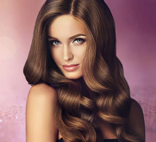 Portret kobiety z długimi, brązowymi, falowanymi włosami, stylizowanymi we Włocławku, na rozmytym, różowo-fioletowym tle z efektem brokatu.