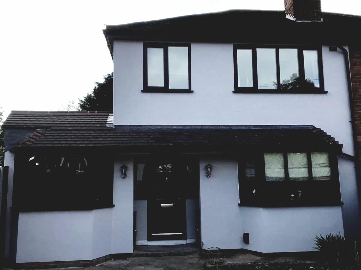 Dwupiętrowy dom z jasnoszarą elewacją, ciemnymi ramami okiennymi i brązowym dachem, sfotografowany w pochmurny dzień.