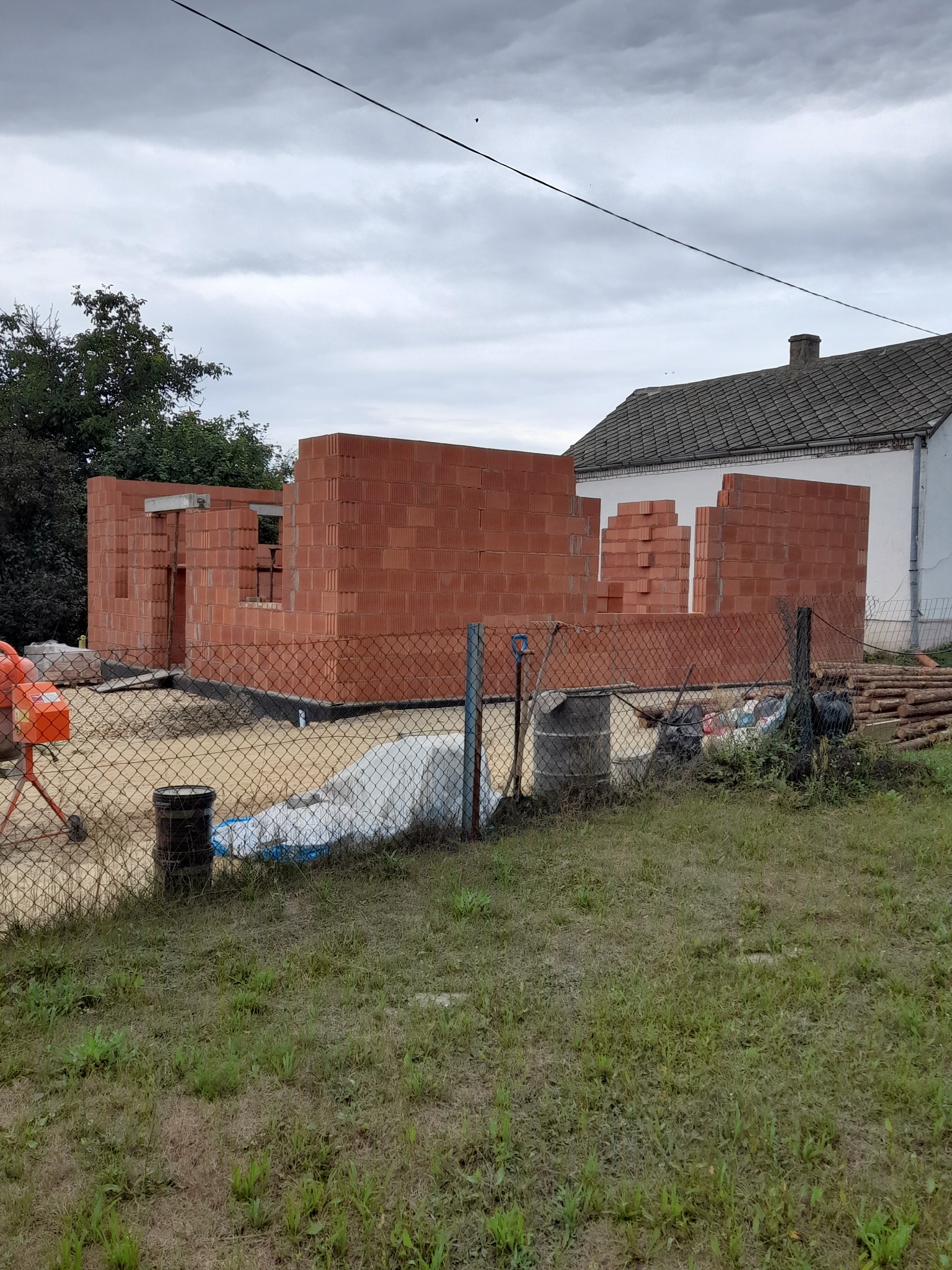 Wznoszone ściany domu z czerwonej cegły ceramicznej, widoczna betoniarka, teren budowy ogrodzony siatką.