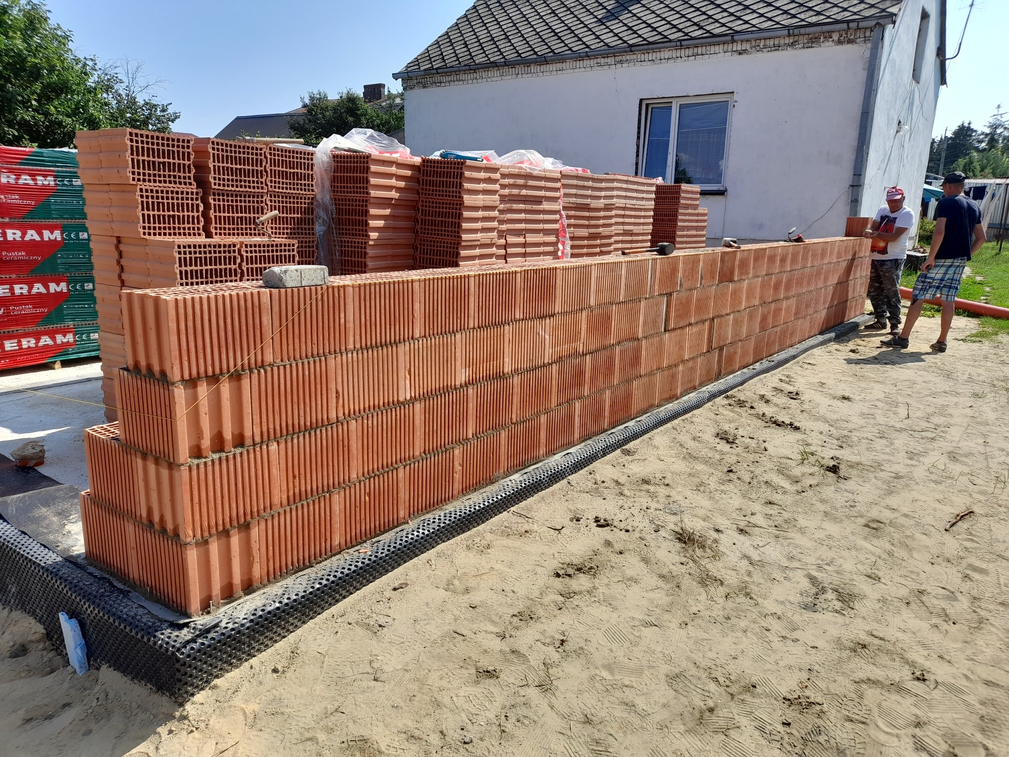 Wznoszenie ściany z czerwonych, porowatych pustaków ceramicznych na fundamencie z folią kubełkową; w tle palety z zapakowanymi pustakami i fragment białego budynku z oknem, dwóch pracowników w tle.