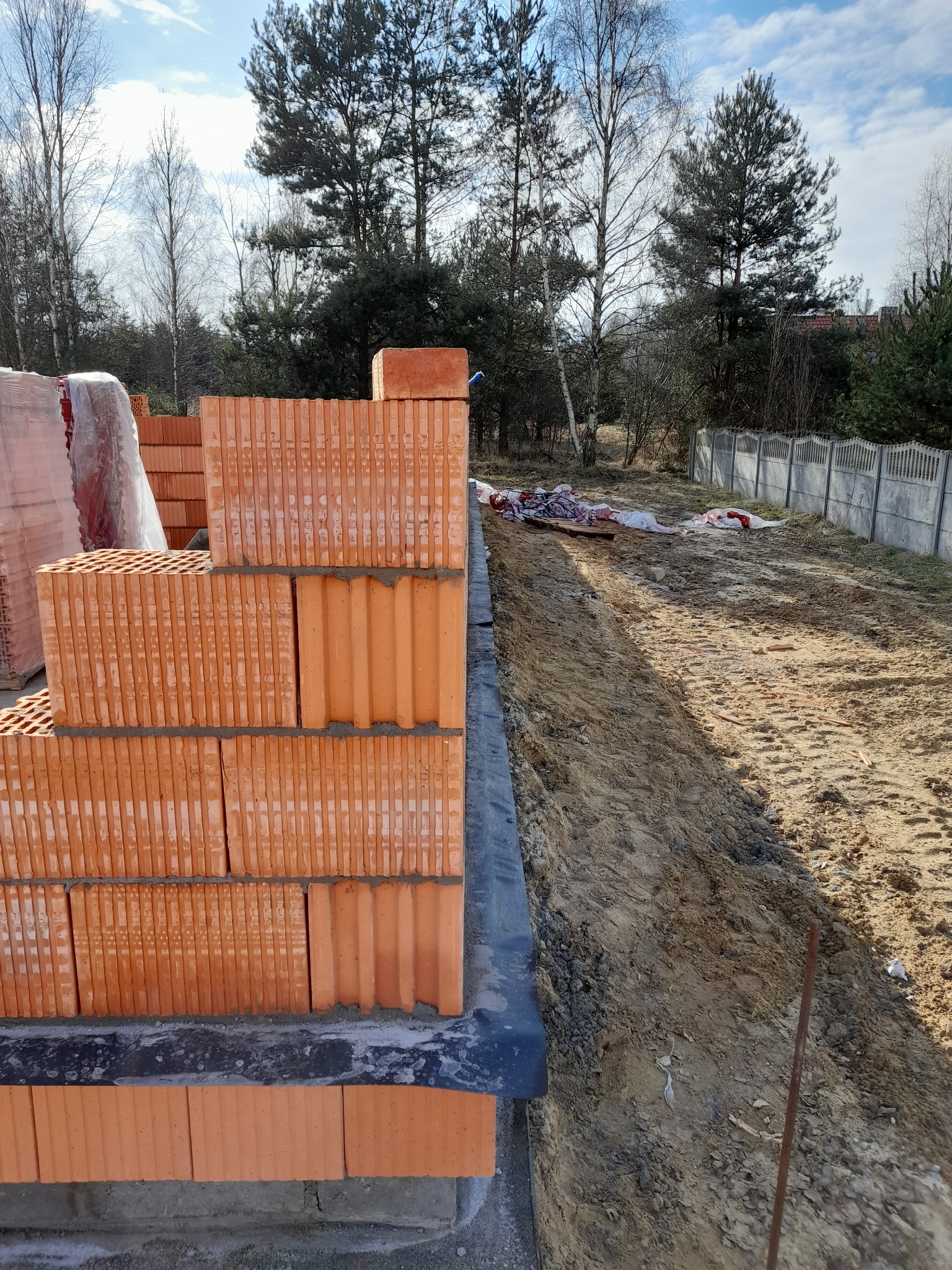 Wznoszenie muru z czerwonej ceramiki poryzowanej na fundamencie w Olsztynie, widoczne materiały budowlane i przygotowany teren pod budowę.