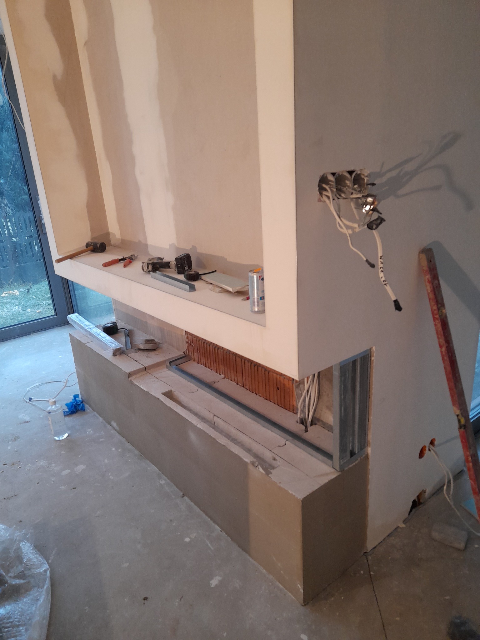 Wnętrze w trakcie prac wykończeniowych: zabudowa kominka z widoczną konstrukcją metalową, cegłami szamotowymi i przewodami elektrycznymi, narzędzia na półce, surowa ściana z puszkami elektrycznymi.