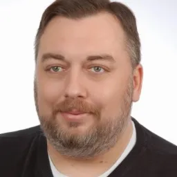 Portret mężczyzny z brodą i zielonymi oczami, ubrany w czarny sweter na jasnym tle.